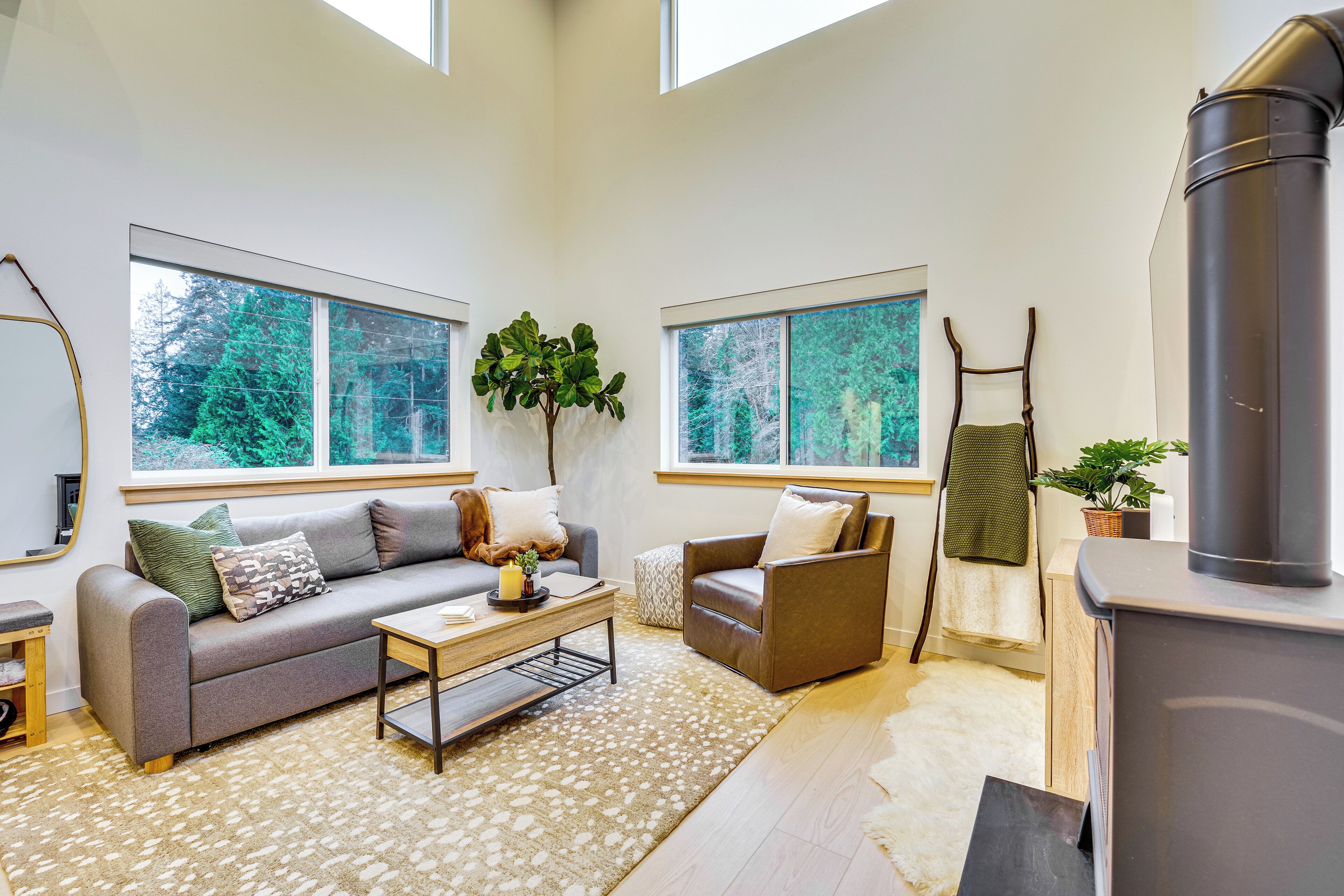 Un salon moderne avec un canapé, un fauteuil et une table basse, de grandes fenêtres donnant sur la verdure et un poêle à bois.
