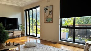 A spacious living room with a large TV, wooden flooring, a white rug, and large windows offering a view of greenery and buildings. - Lipscombe Mt Wellington view luxury house (HOBART)