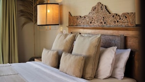 A hotel room with a bed, pillows, a headboard with intricate carvings, a wall-mounted lamp, and a nightstand. - Alea Estate by Villa Finder (Kerobokan)