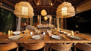 A dining area with a wooden table set for a meal, surrounded by chairs, with a sofa and a dining table in the background. - Alea Estate by Villa Finder (Kerobokan)