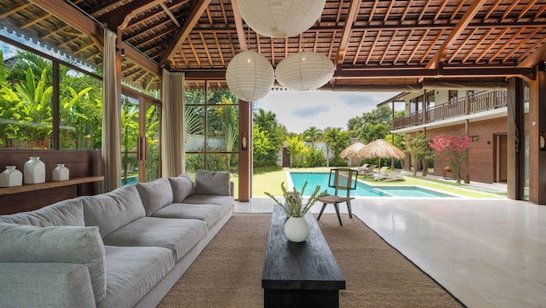 A spacious living area with a large sofa, a coffee table, and a view of a swimming pool and lush garden. - Alea Estate by Villa Finder (Kerobokan)