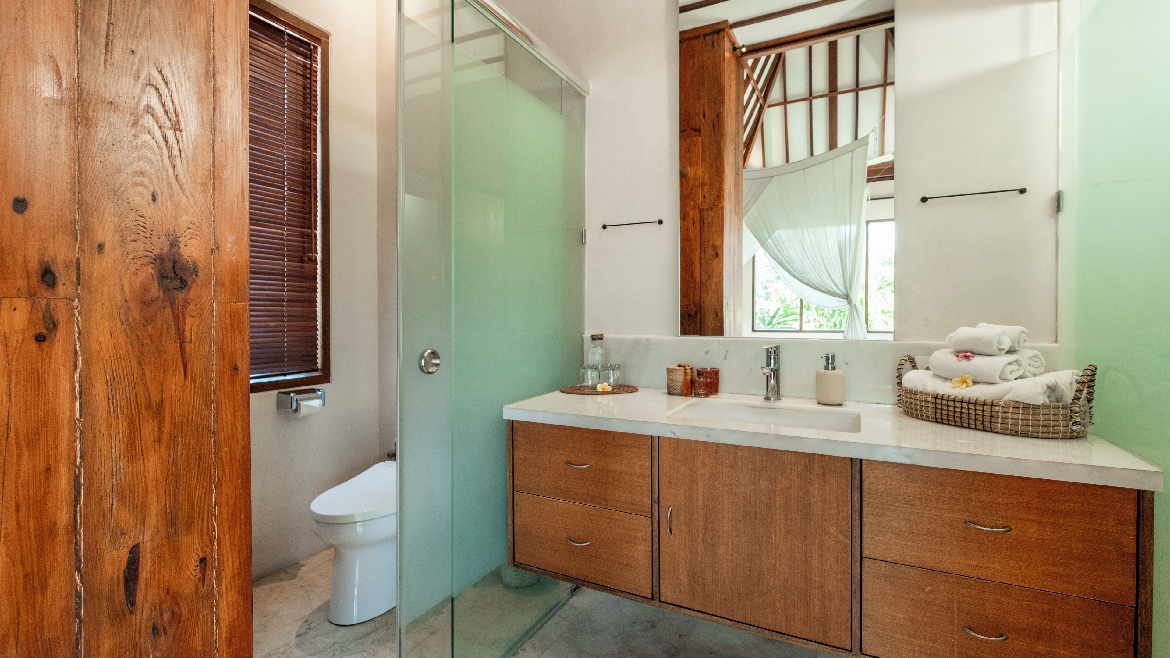 A bathroom with a wooden door, a toilet, a glass shower enclosure, a vanity with a mirror, and a window with a view of greenery.