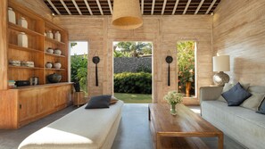 A modern living room with a sofa, a coffee table, and a bookshelf. There is a view of a garden through a doorway. - Alea Estate by Villa Finder (Kerobokan)
