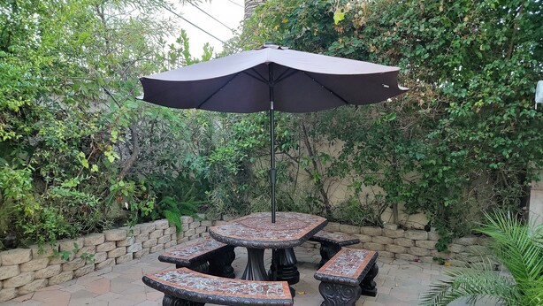An outdoor patio with a table and benches, surrounded by a stone wall and lush greenery.