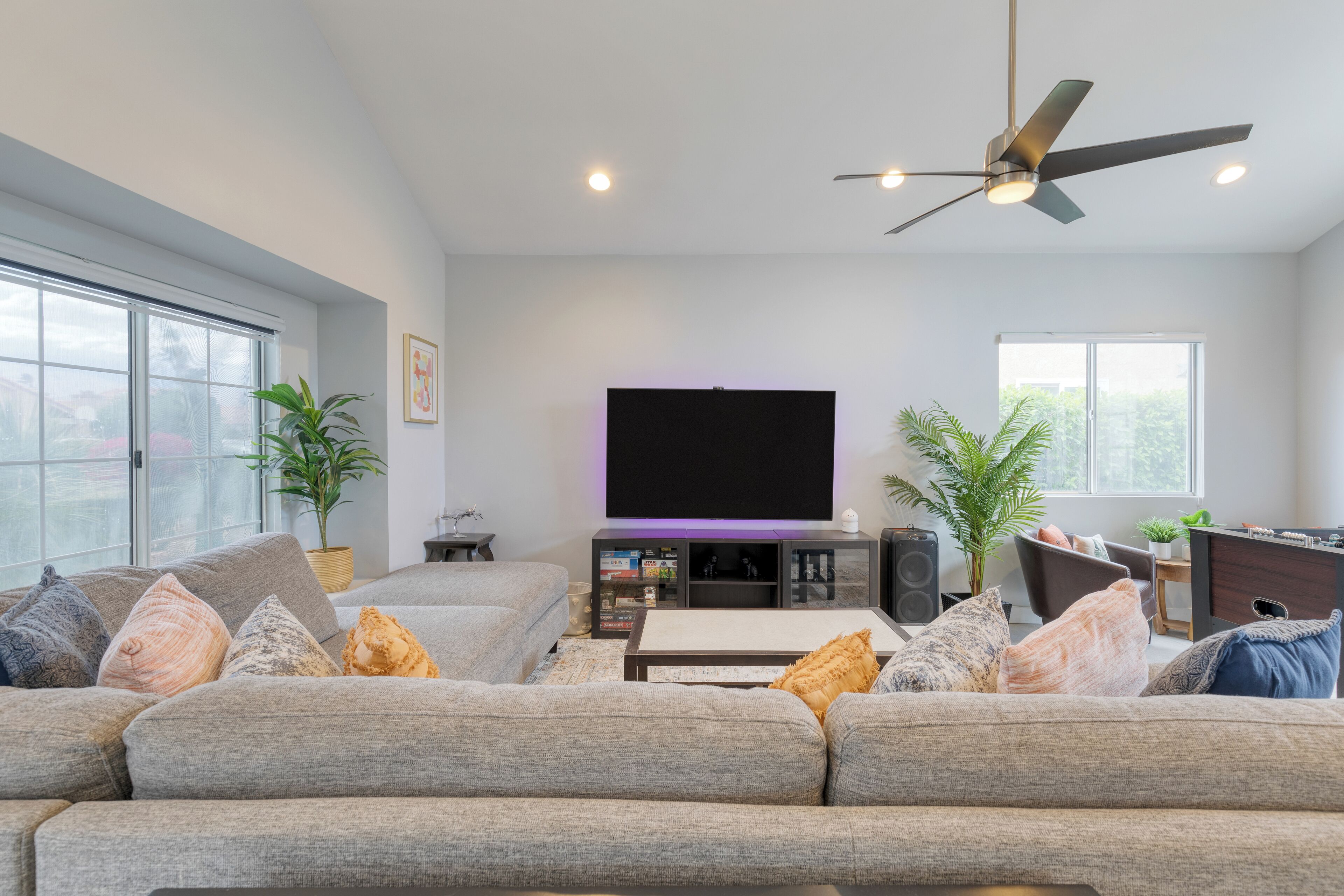 A modern living room with a large flat-screen TV, a ceiling fan, and a comfortable seating area with multiple sofas and cushions.