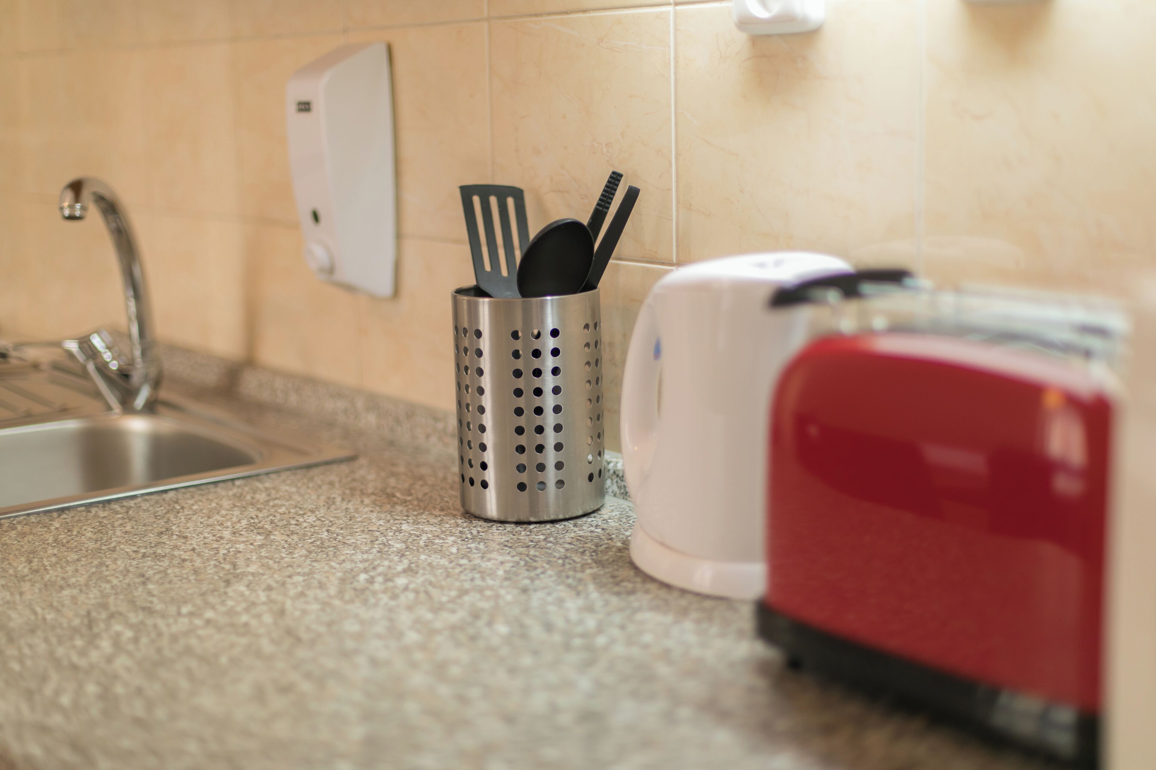 A kitchen with a stainless steel sink, a perforated metal container holding kitchen utensils, and a red toaster.