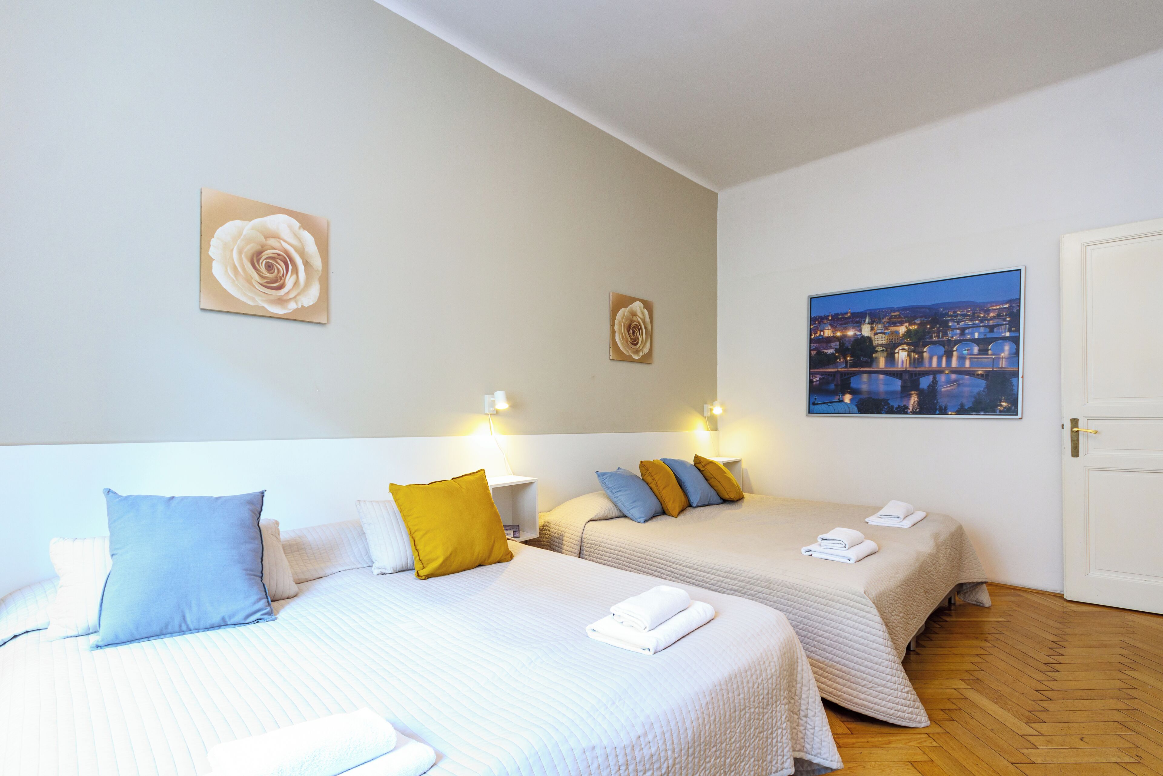 A hotel room with two beds, each with a white bedspread and pillows in blue, yellow, and white. There are wall-mounted lights and a framed picture of a cityscape.