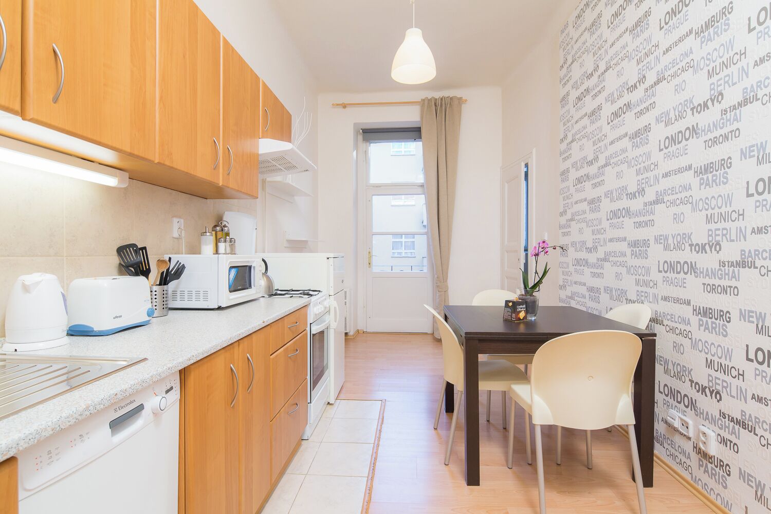 A compact kitchen with wooden cabinets, a microwave, a toaster, and a sink. There's a dining area with a table and chairs, and a wall with city names.