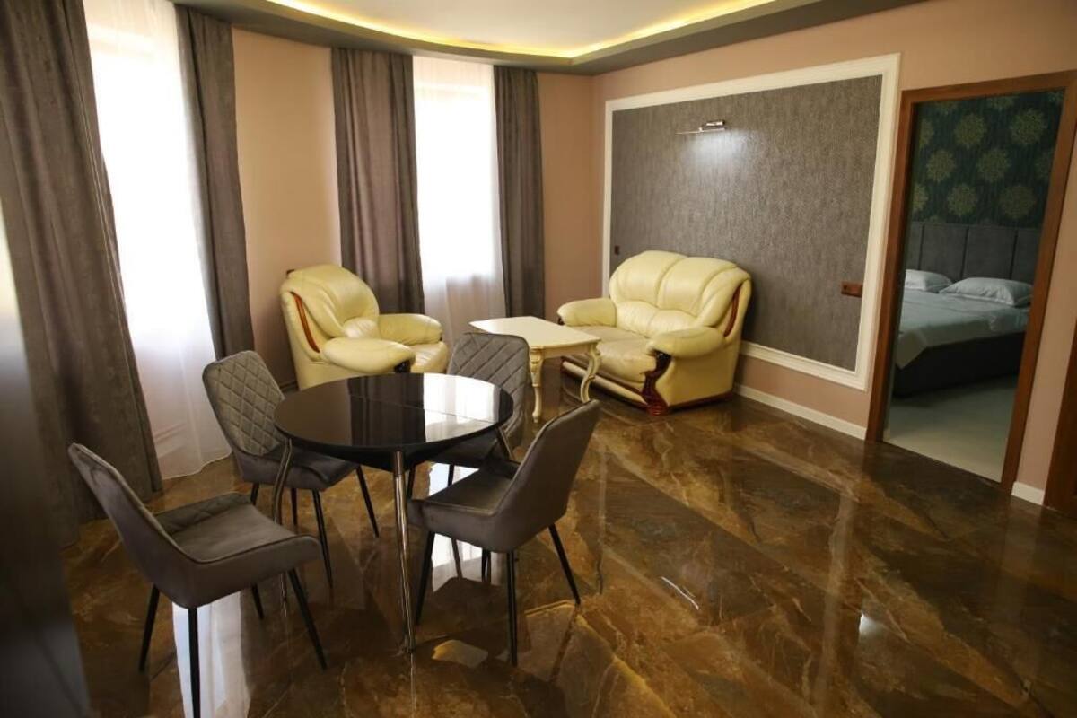 A room with a round table, four chairs, two sofas, a bed, and a large window with curtains.