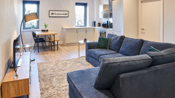 A modern living room with a grey sofa, a wooden coffee table, and a floor lamp. There is a dining area with a table and chairs, and a kitchen with white cabinets and a stove. - Host Stay Mariposa Waterloo (Liverpool)