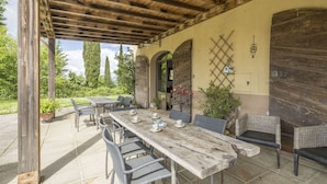 An outdoor patio with a rustic wooden table and chairs, surrounded by greenery and a stone floor.