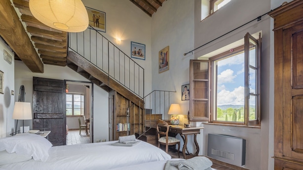 A spacious bedroom with two beds, a wooden staircase, a desk with a lamp, and a large window with a view of greenery.