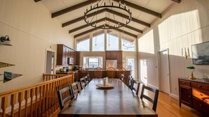 Uma ampla sala de jantar com uma longa mesa de madeira, cadeiras de madeira e um candelabro. Cozinha ao fundo com armários de madeira e um micro-ondas.