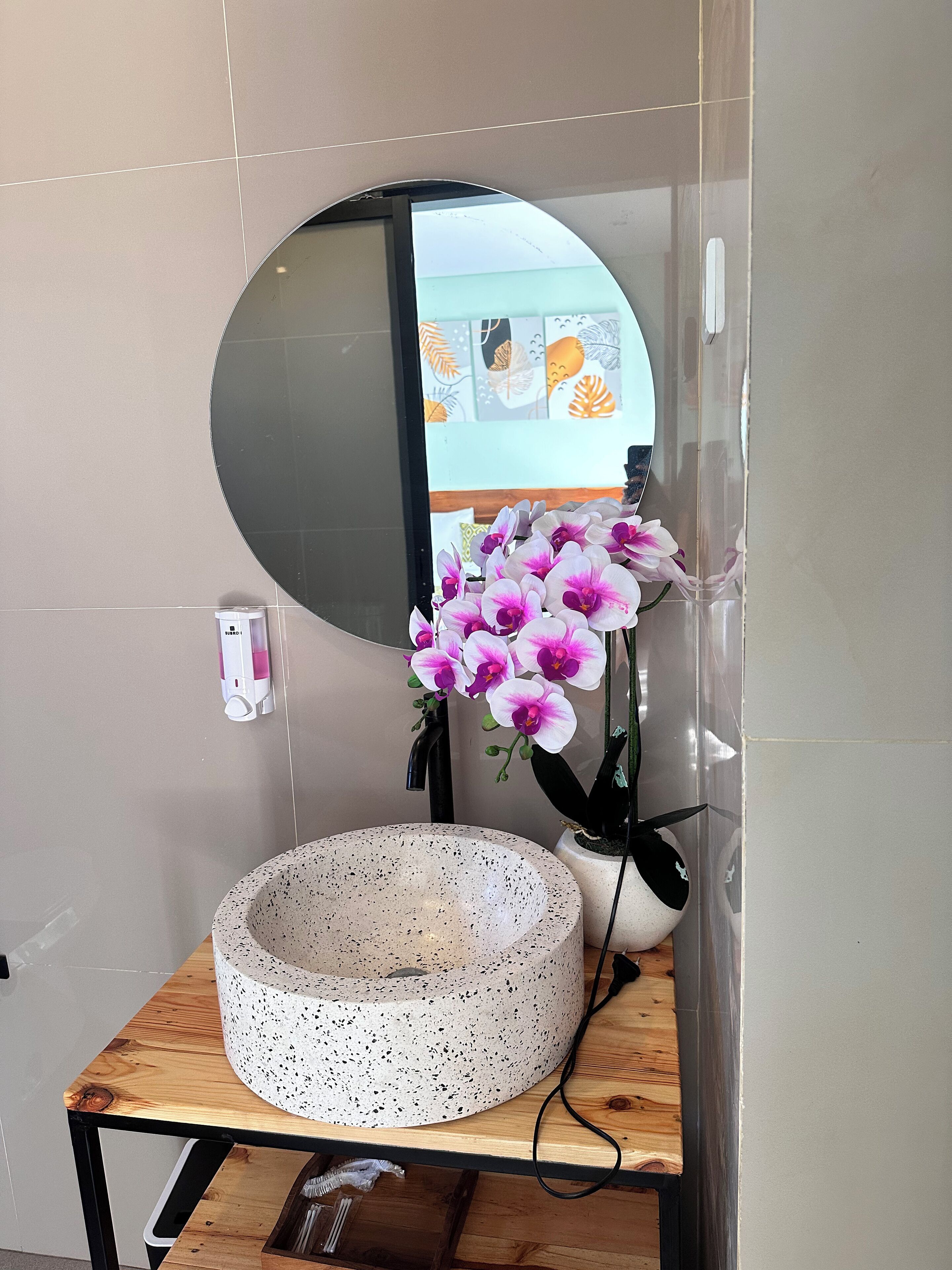 Un lavamanos de baño con un espejo redondo, un lavabo de cerámica con motas y un jarrón con orquídeas rosadas.