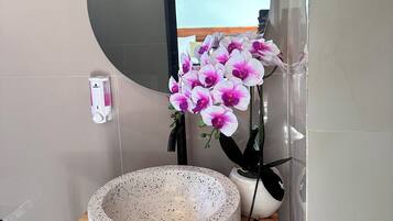 Un lavamanos de baño con un espejo redondo, un lavabo de cerámica con motas y un jarrón con orquídeas rosadas.