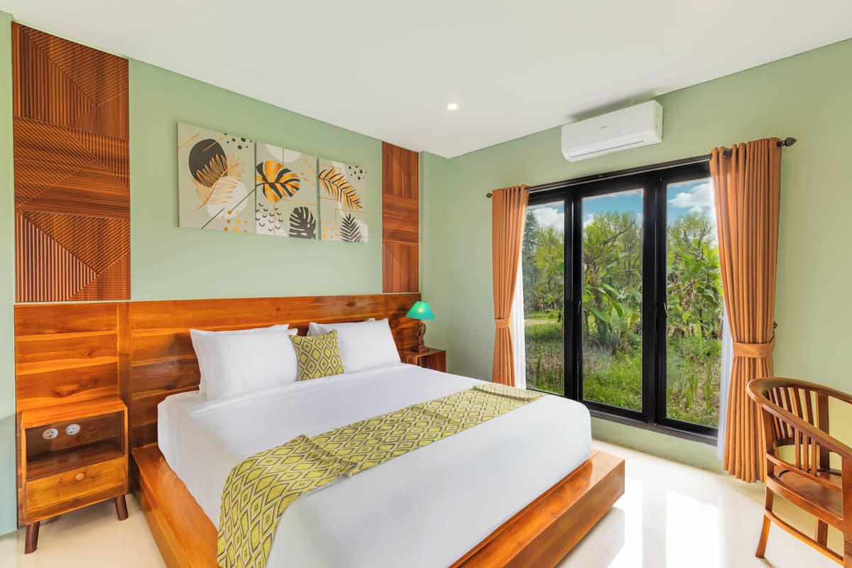 A bedroom with a wooden headboard, a bed with white linens, a green patterned throw, a nightstand with a lamp, and a view of greenery through the window.