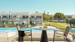 Un balcon avec une piscine, deux fauteuils en osier, une table ronde avec des fruits, et un bâtiment moderne en arrière-plan.