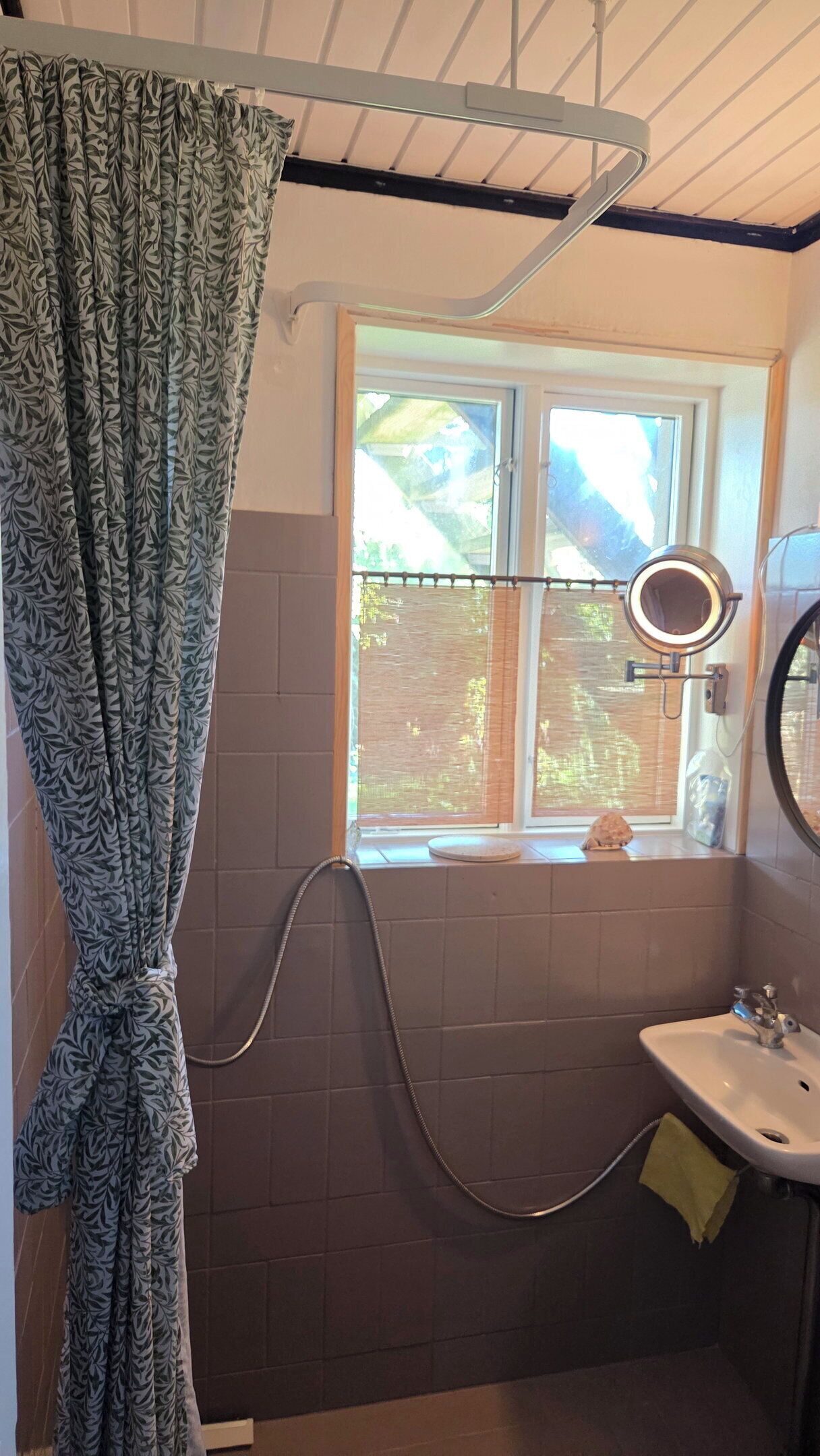 A bathroom with a shower, a window with blinds, a sink, and a mirror.