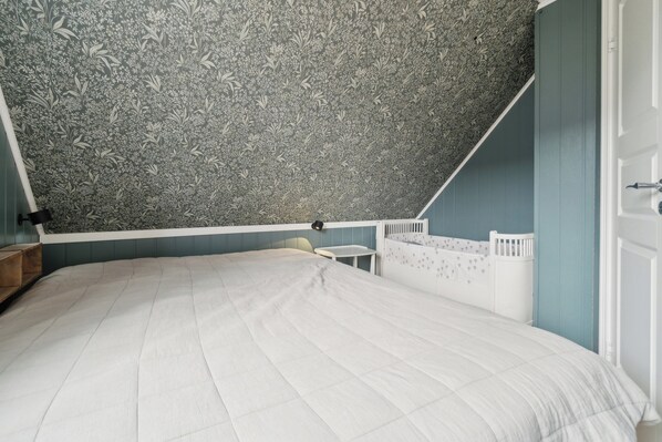 Ein Schlafzimmer mit schrägem Dach, einem weißen Bett, einem kleinen Tisch und einem weißen Wiege.