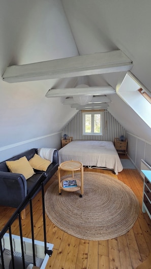 A loft bedroom with a sloped ceiling, a bed, a sofa, a small wooden table, and a window.
