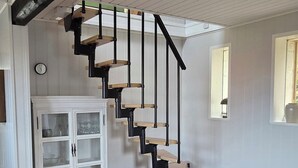 Eine Treppe mit schwarzer Handlauf und Holzstufen, ein weißer Schrank mit Glastüren, ein Korbwägelchen, ein gestreiftes Sofa und ein runder Holztisch.