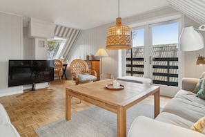 A modern living room with a TV, wooden furniture, a dining table, and a large window with a view.