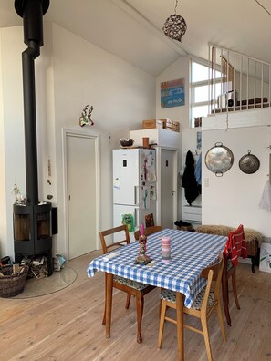 Un coin repas avec une nappe à carreaux, des chaises en bois, un poêle en bois noir, un réfrigérateur blanc et un escalier menant à un niveau supérieur.