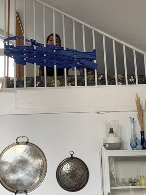 Un escalier avec des rampes blanches, une personne qui jette un coup d’œil derrière un rideau bleu, et une étagère avec de la verrerie et un vase décoratif.