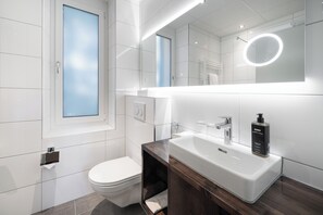 A modern bathroom with a white sink, a mirror with a circular light, and a toilet.