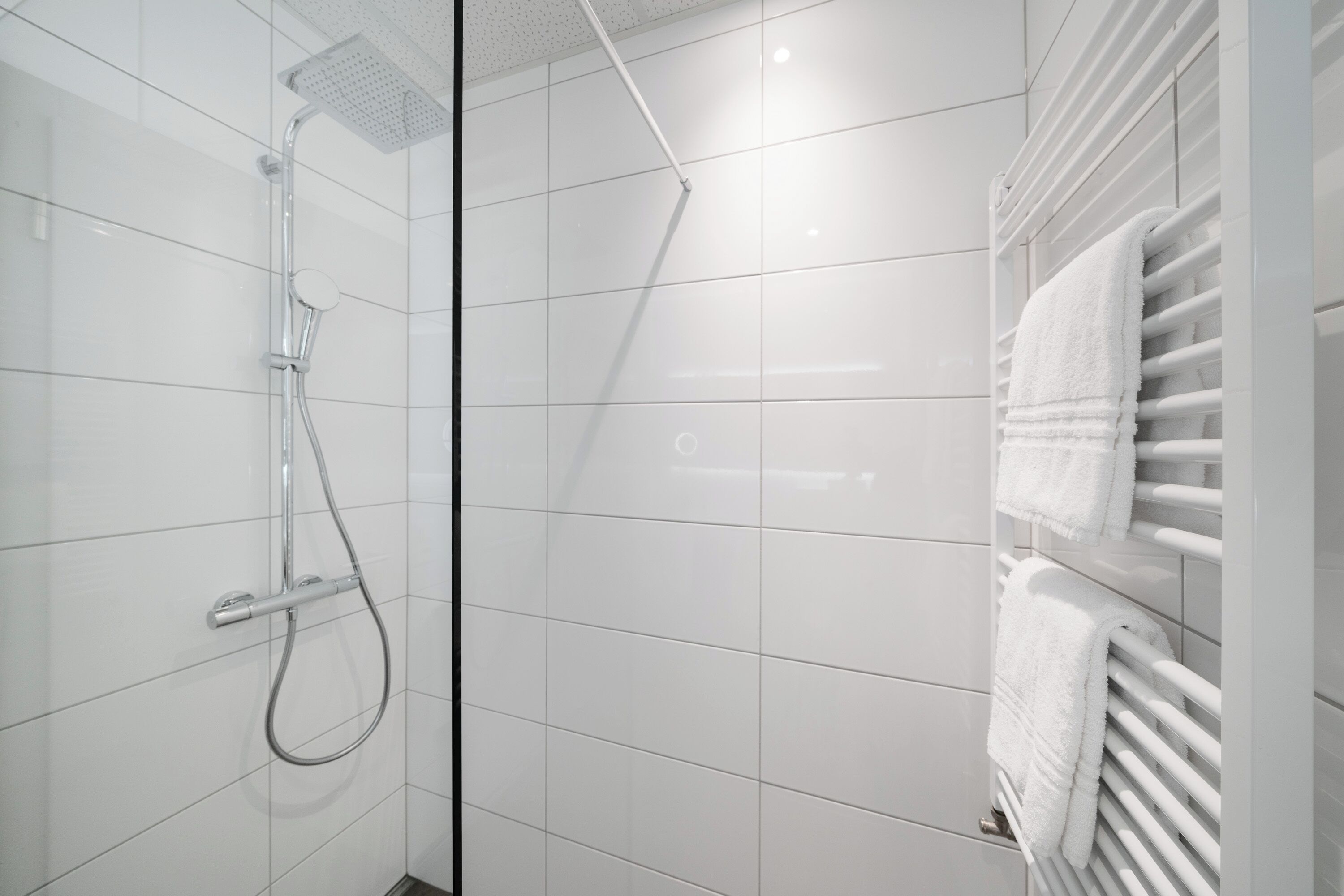 Une salle de bain moderne avec des murs carrelés de blanc, une douche équipée d’un pommeau de douche à main et un porte-serviettes avec deux serviettes blanches.