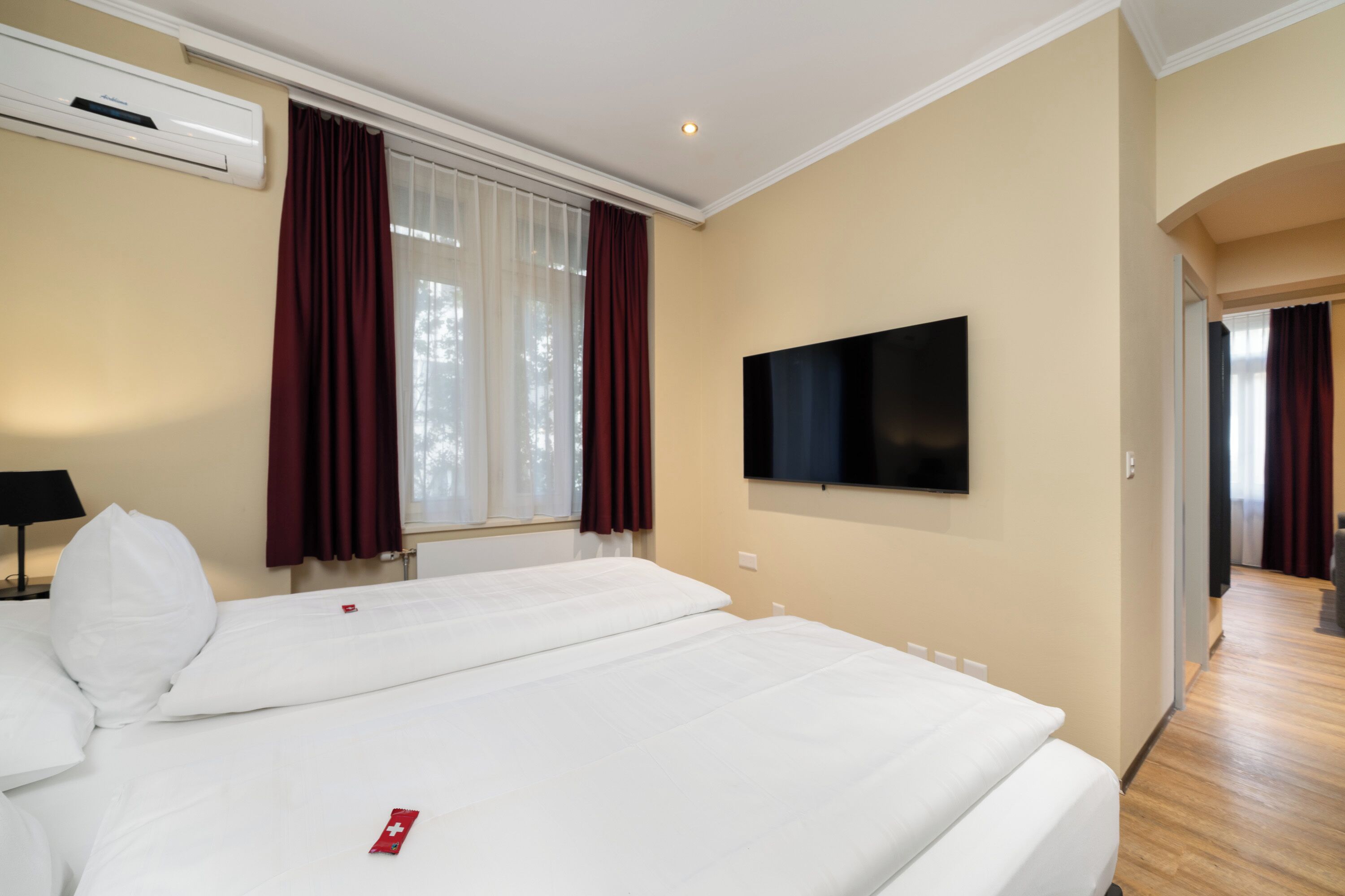 Ein Hotelzimmer mit einem großen Bett, einem Flachbildfernseher, einer Klimaanlage und einem Vorhangfenster.