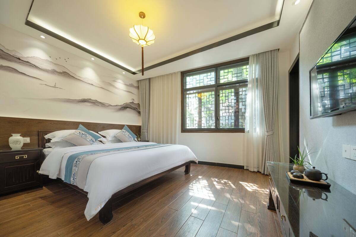 Un dormitorio moderno con una cama grande, suelo de madera, una pared decorativa, una ventana con cortinas y un televisor montado.