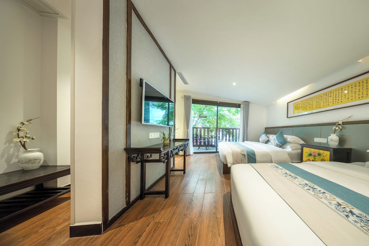 Habitación de hotel con cama, televisión, escritorio, planta y balcón con vista a árboles.