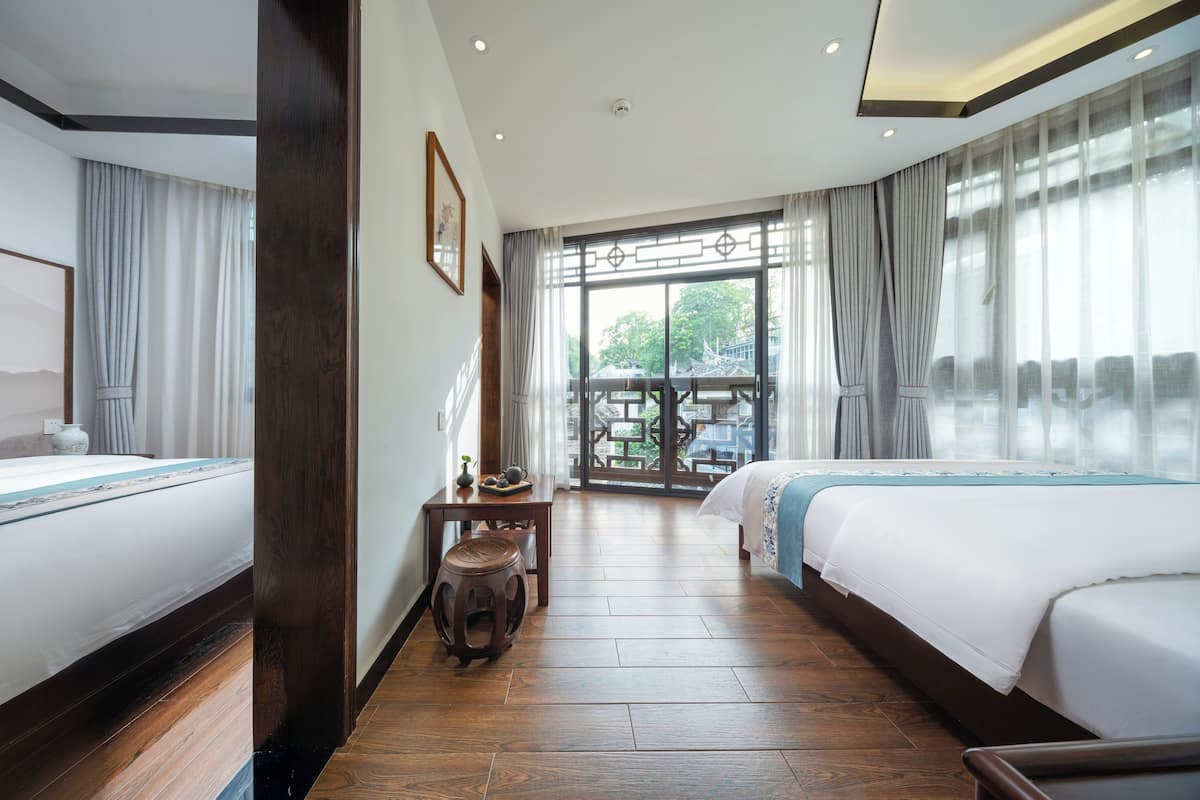 Habitación de hotel con dos camas, piso de madera, una mesita con tetera y vistas a un edificio tradicional a través de amplios ventanales.