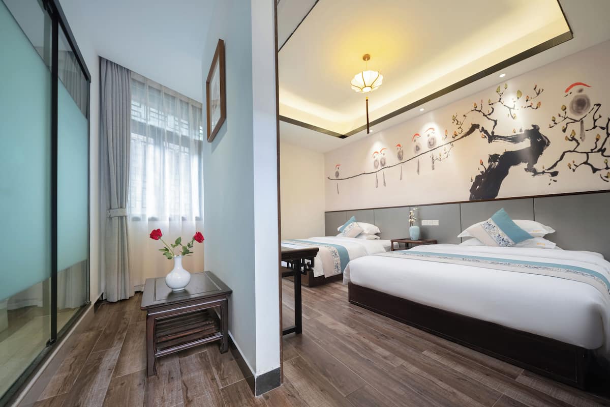 Una habitación de hotel moderna con una cama grande, una cama más pequeña, una mesita de noche con un jarrón de flores, una obra de arte en la pared y una lámpara en el techo.