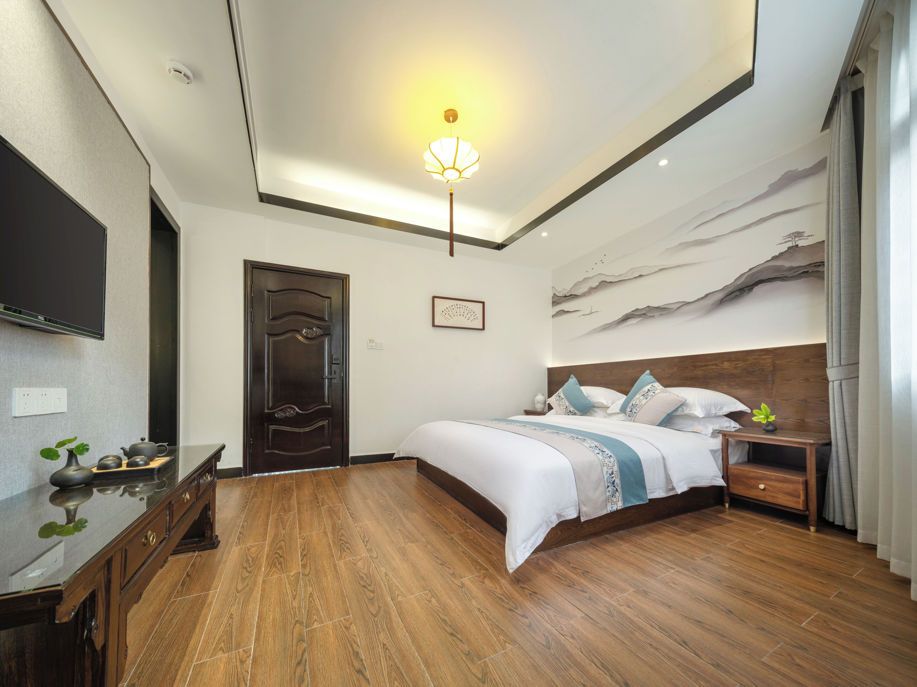 A modern bedroom with a wooden floor, a bed with white bedding, a nightstand, a TV mounted on the wall, and a ceiling light fixture.