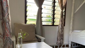 A room with a white bed, a small white table with a bottle and a plant, and a patterned curtain. - MUMBI VILLA WATAMU (Watamu)