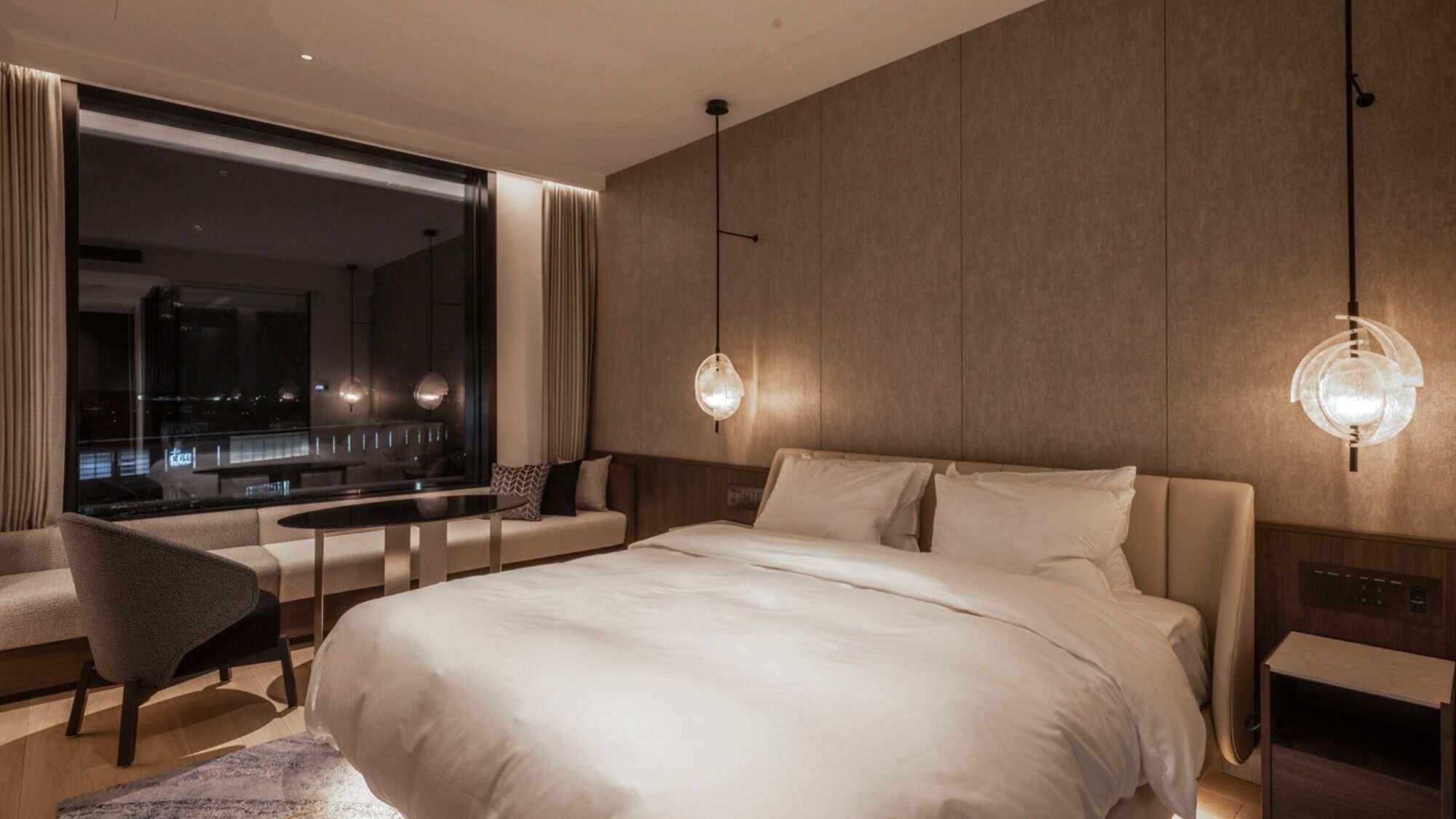 Habitació d'hotel moderna amb un llit gran, zona de seients, un finestral gran amb vista a la ciutat i dues làmpades penjants.