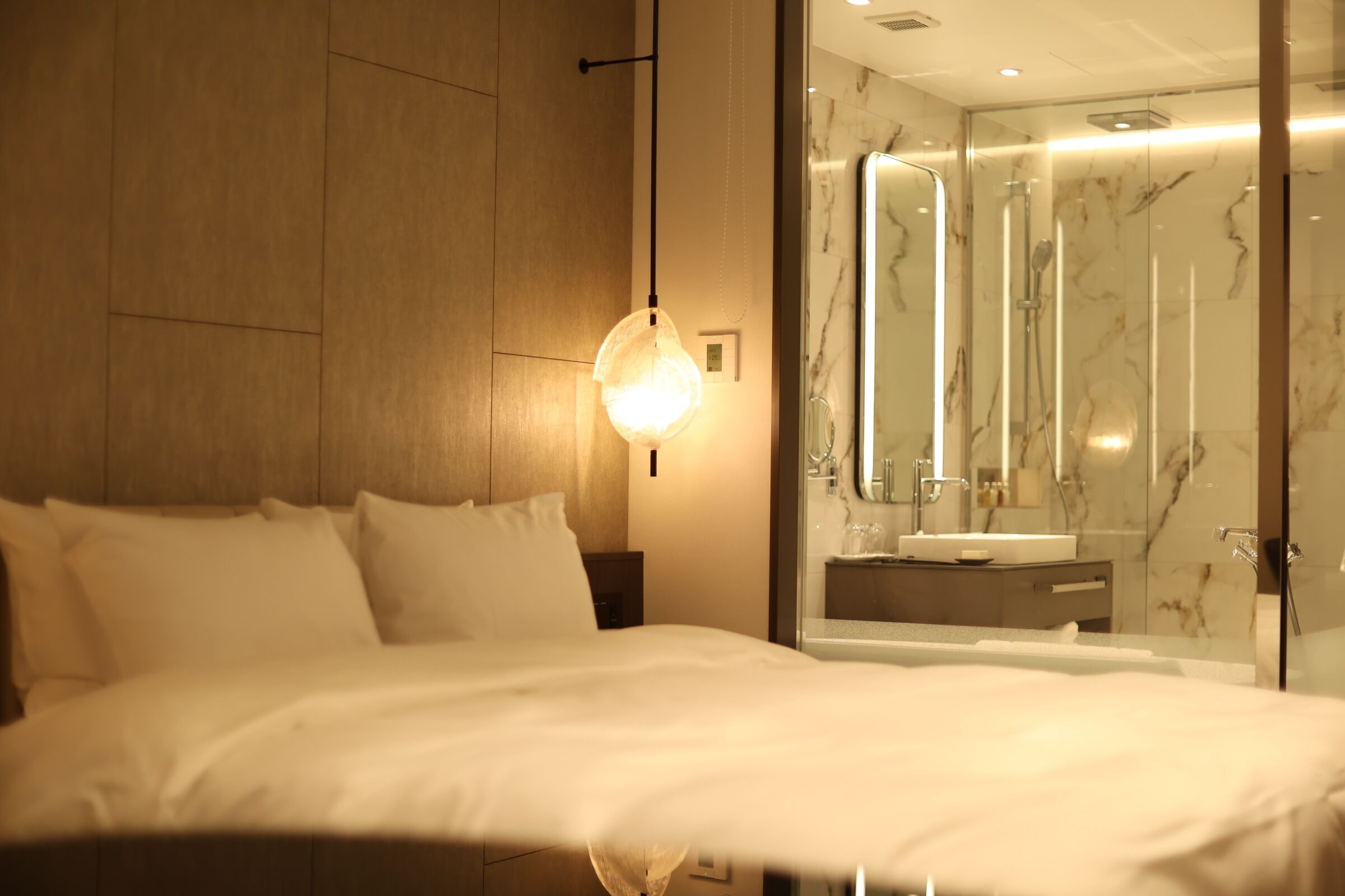 Une chambre d’hôtel comprenant un lit, une lampe de chevet, une cabine de douche en verre et un mur recouvert de carreaux de marbre.