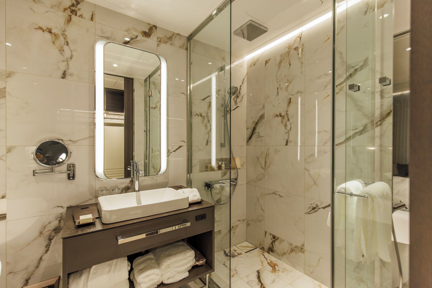 Un baño moderno con ducha envuelta en vidrio, pared de azulejos de mármol, espejo redondo y tocador con lavamanos y espacio para toallas.