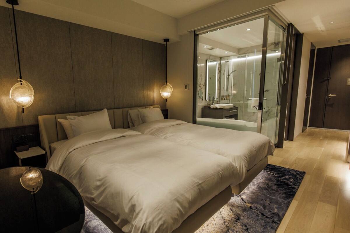 A modern hotel room with a large bed, bedside tables, a round black table, and a glass-enclosed bathroom.