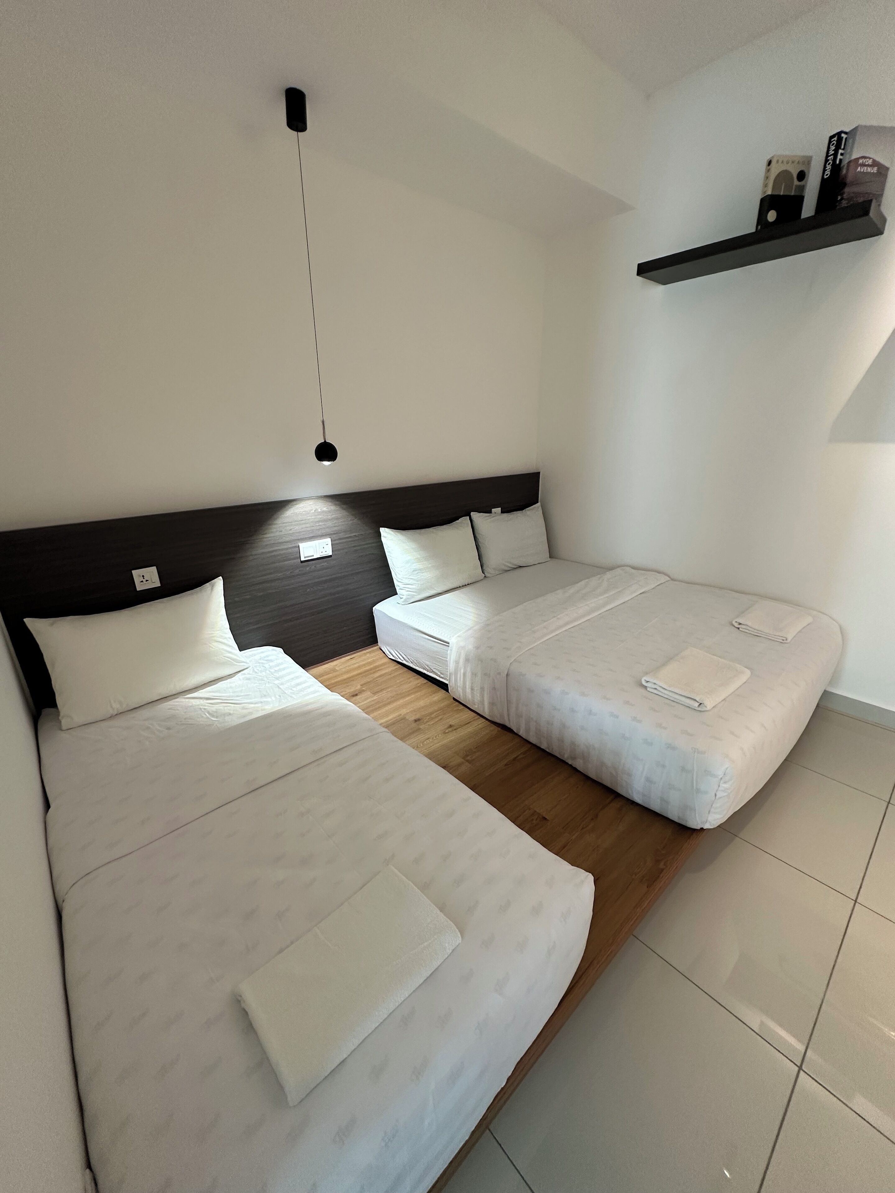 Moderni hotellihuone, jossa on kaksi sänkyä, tumma pääty sekä pieni hylly, jossa on muutamia esineitä, ja riippuvalaisin.