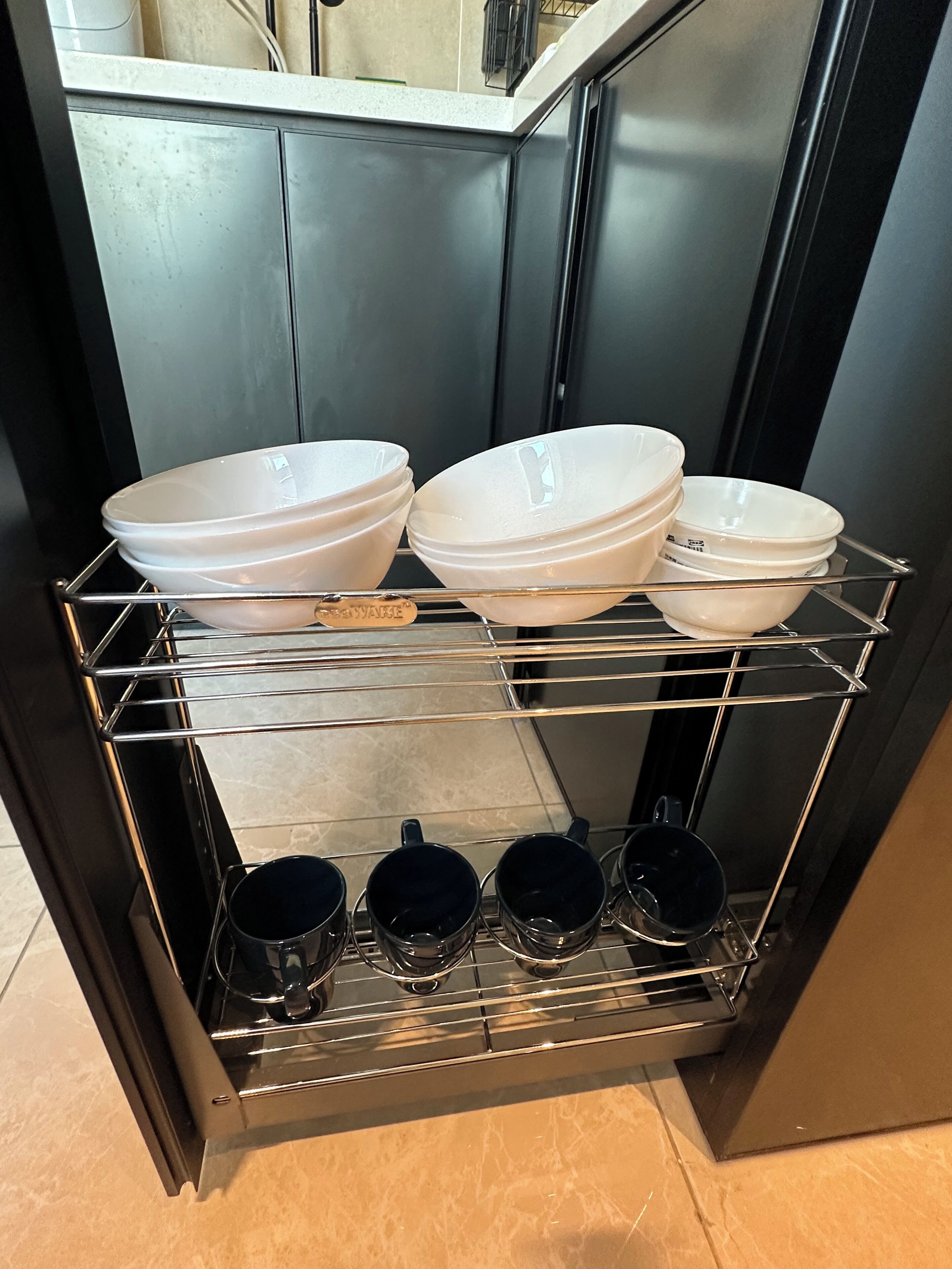 Una cocina con una estantería metálica que contiene cuencos blancos y tazas negras.