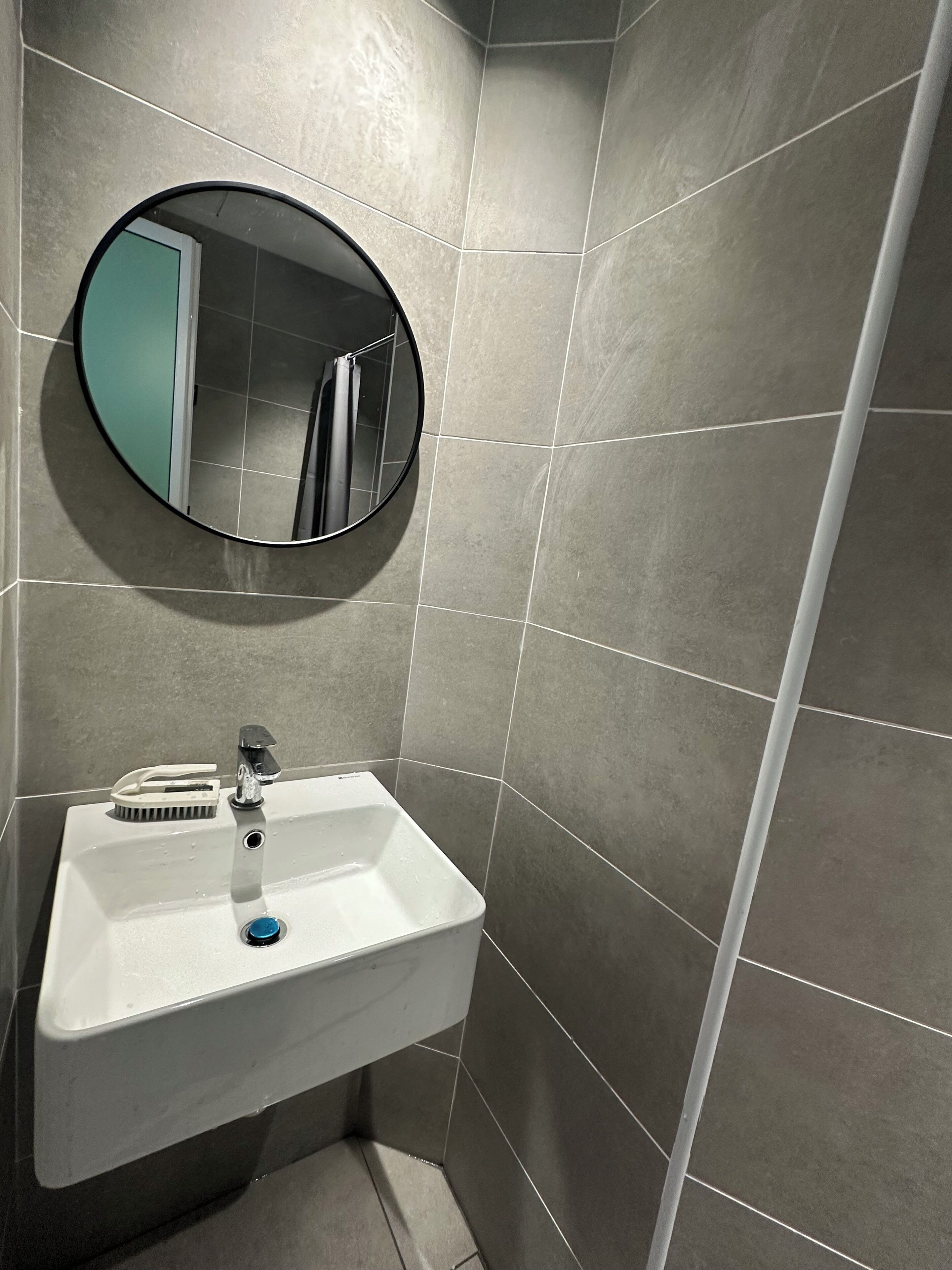 A bathroom with a white sink, a round mirror, and grey tiles.