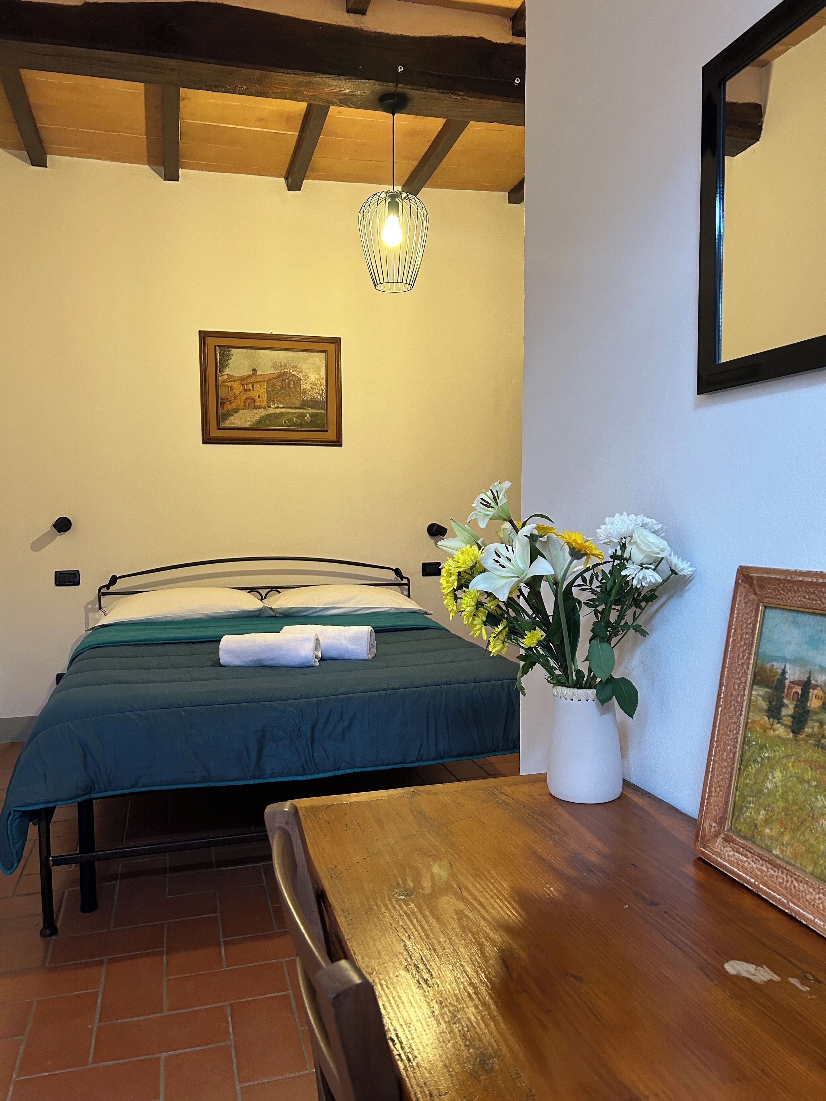 A bedroom with a bed, a wooden table, a vase with flowers, and a painting on the wall.
