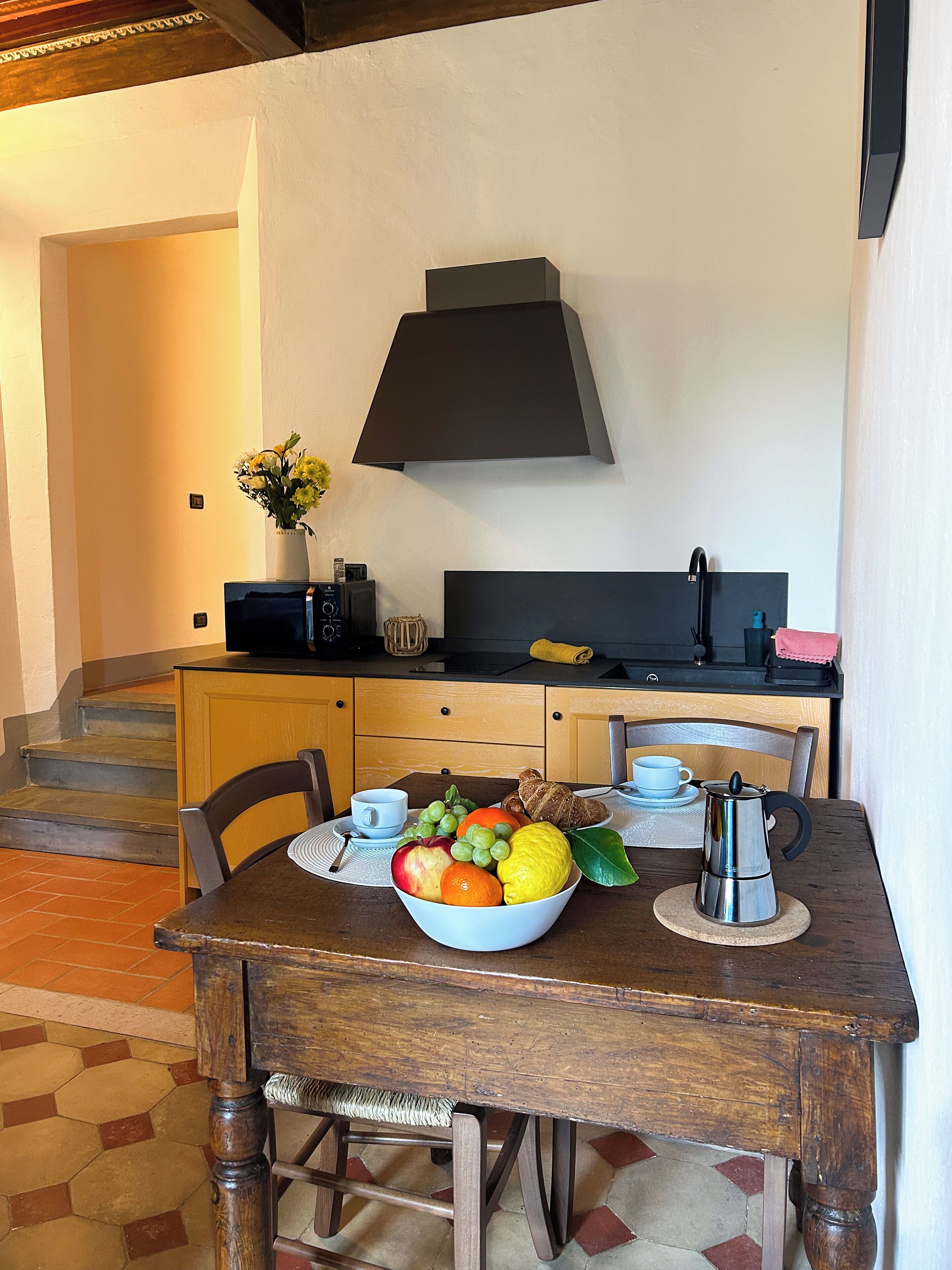 A dining area with a wooden table, chairs, a bowl of fruit, a coffee pot, and a kitchen in the background.