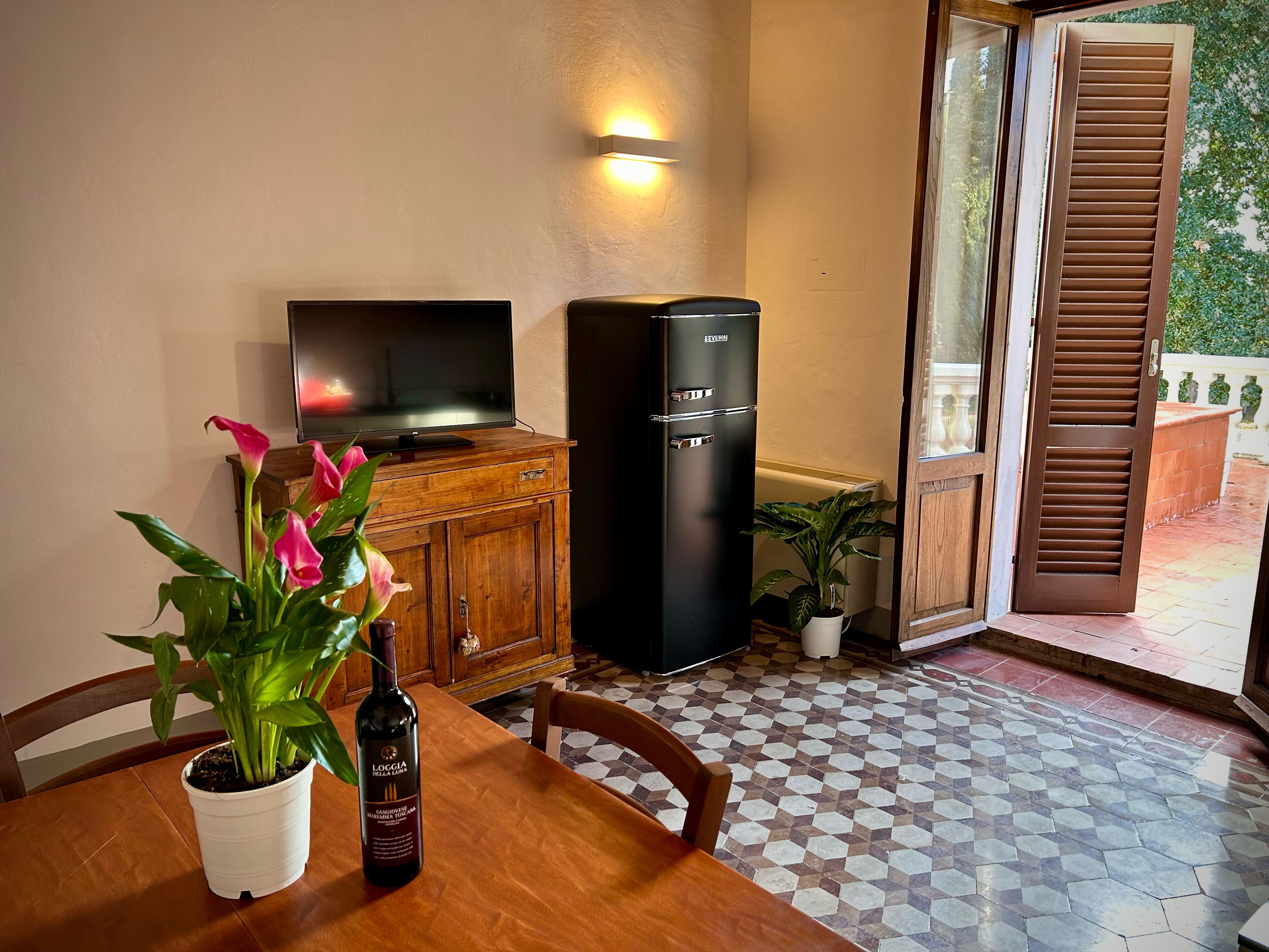 Un comedor con una mesa, sillas, una botella de vino, un televisor, un refrigerador negro y una planta en maceta.