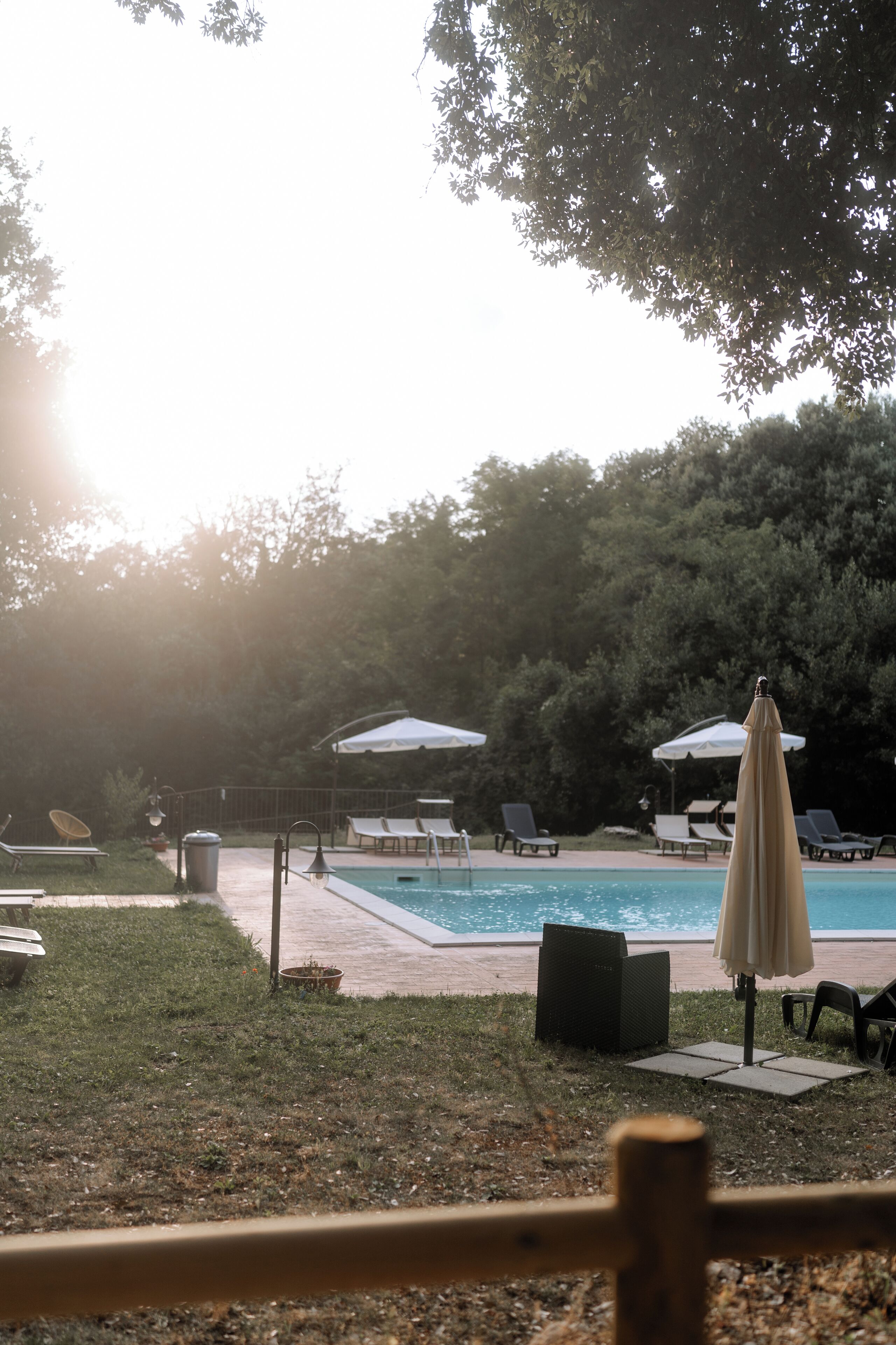 Un área junto a la piscina con sillones de descanso y sombrillas, rodeada de árboles.
