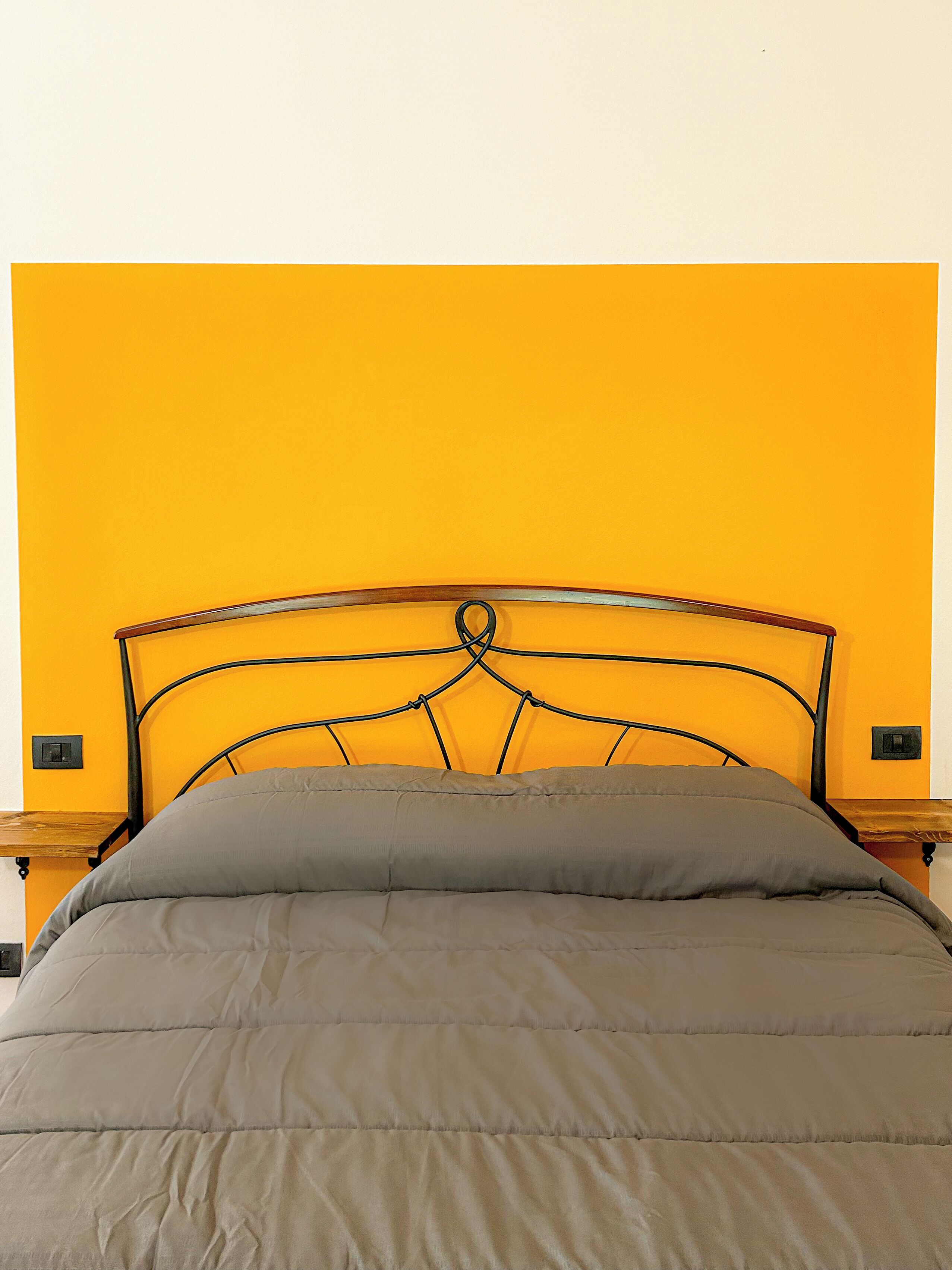 A bed with a metal frame and a grey duvet against a yellow wall.