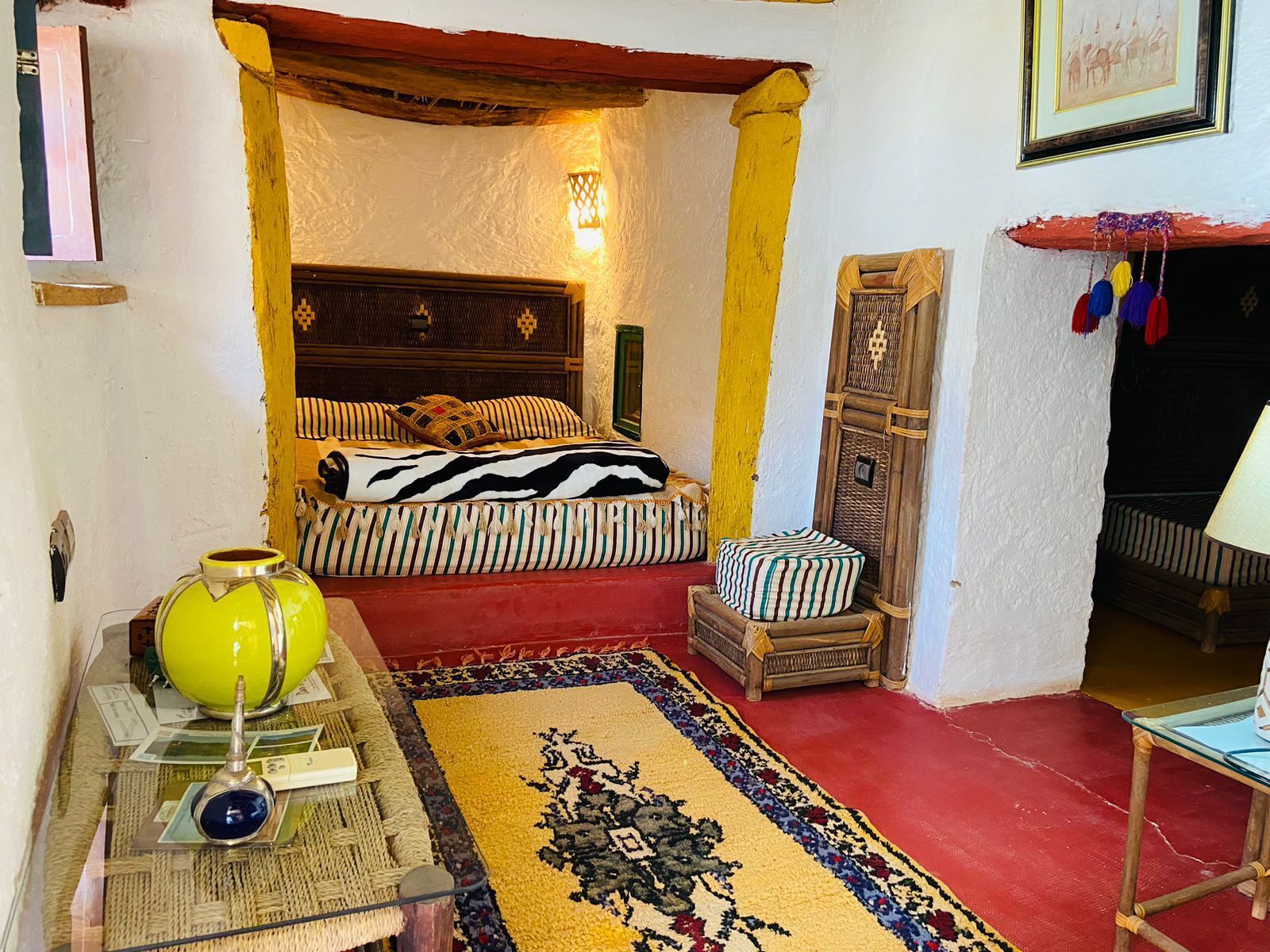 Een kamer met een bed, een klein tafeltje met een gele vaas, een kamerbreed tapijt en een wandplank.